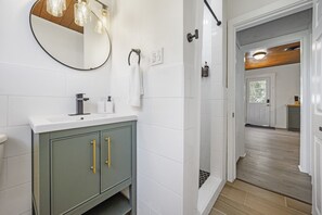 A modern bathroom with a white tiled wall, a green cabinet with gold handles, a round mirror, and a shower area.