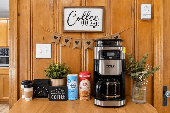 A coffee bar setup with a coffee maker, coffee cups, sugar, creamer, and a sign that reads 'Coffee Bar'.