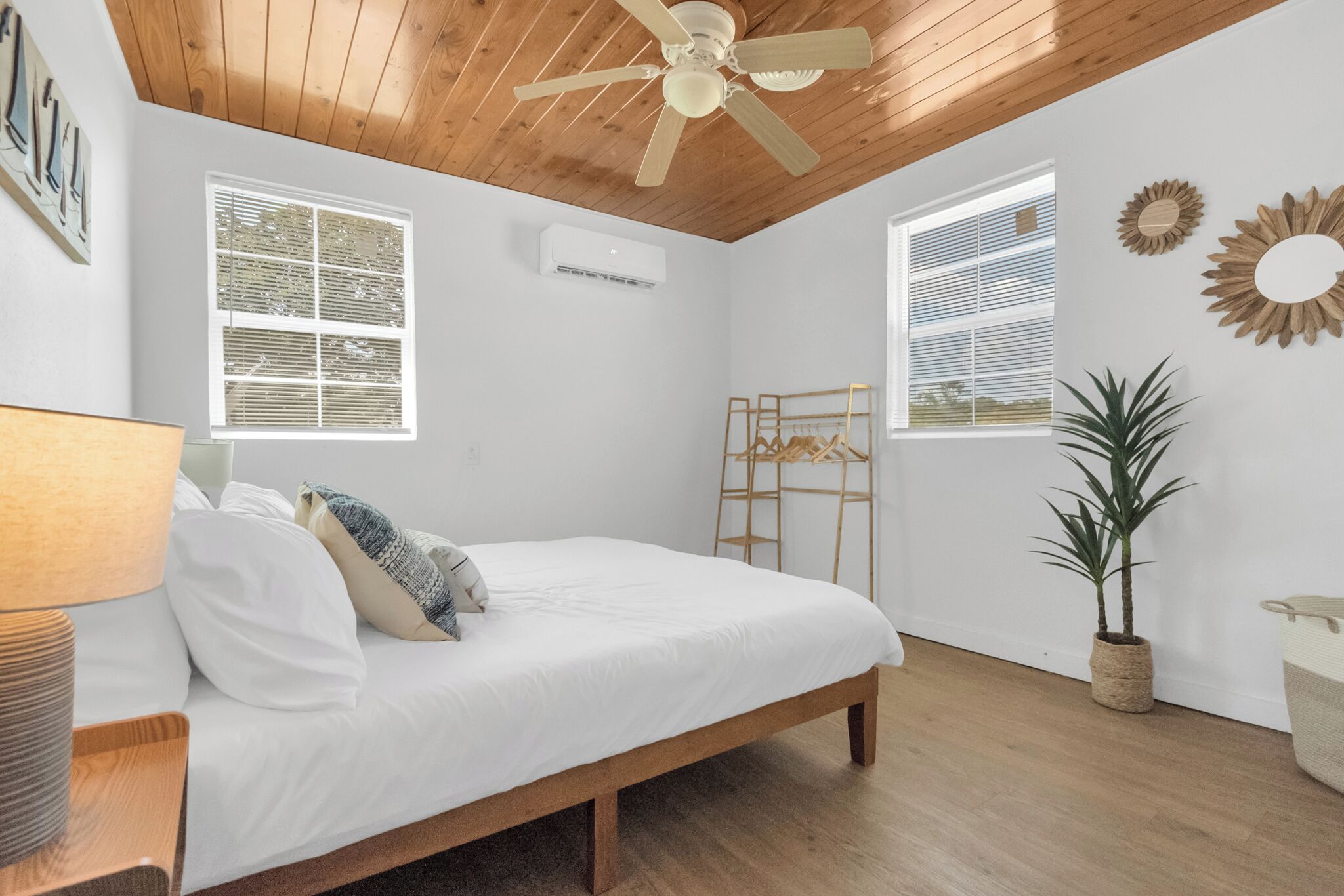 Une chambre avec un plafond en bois, un lit, un portant à vêtements suspendu, une plante en pot et deux fenêtres équipées de stores.