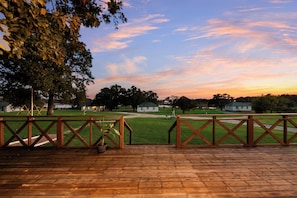 A wooden deck with a railing, overlooking a grassy area with trees and a building in the distance.