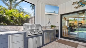 Cocina exterior moderna con una parrilla integrada, armarios de acero inoxidable y un amplio ventanal con vistas a un jardín.