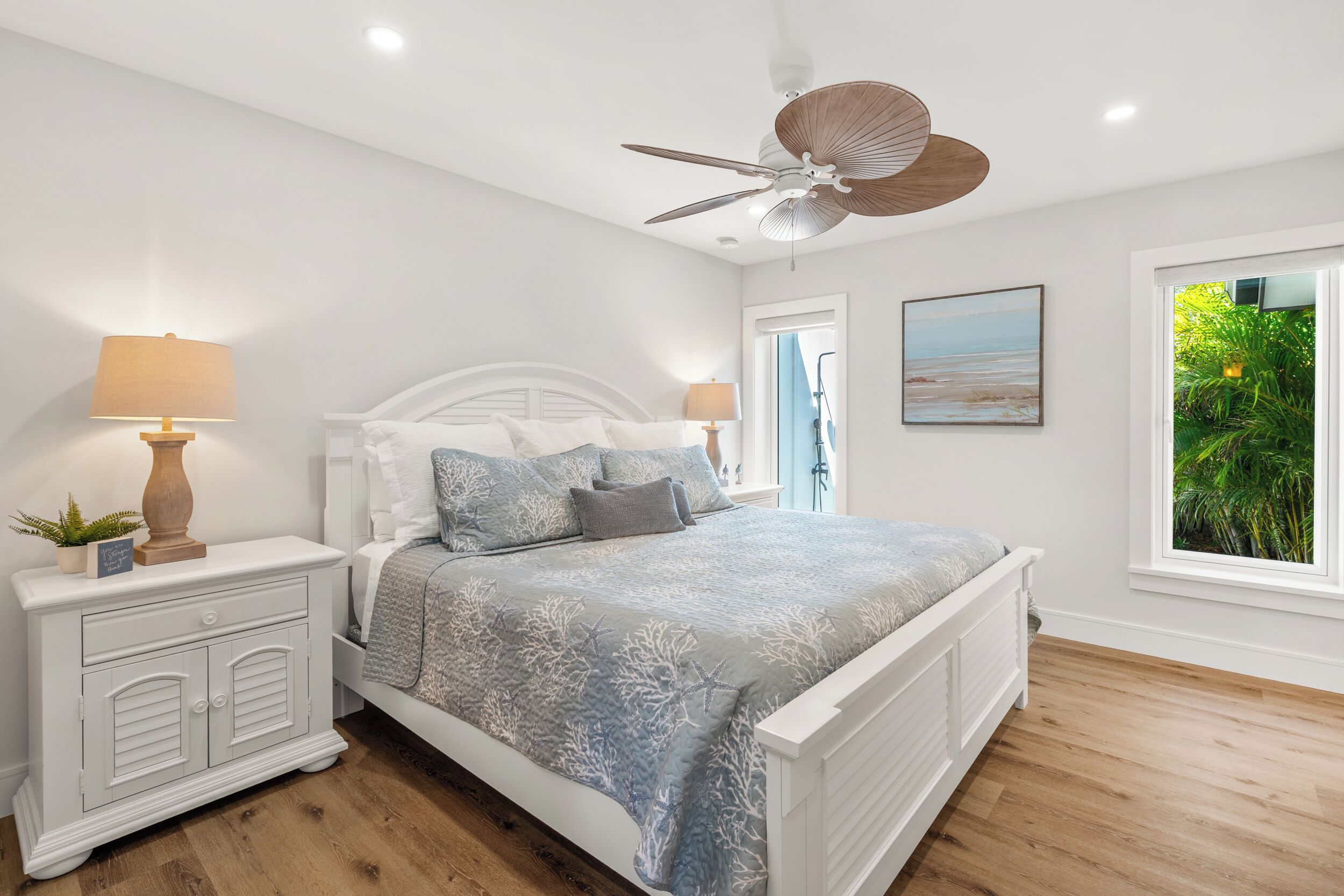 A bedroom with a bed, nightstands, a ceiling fan, a window with a view of greenery, and a painting on the wall.