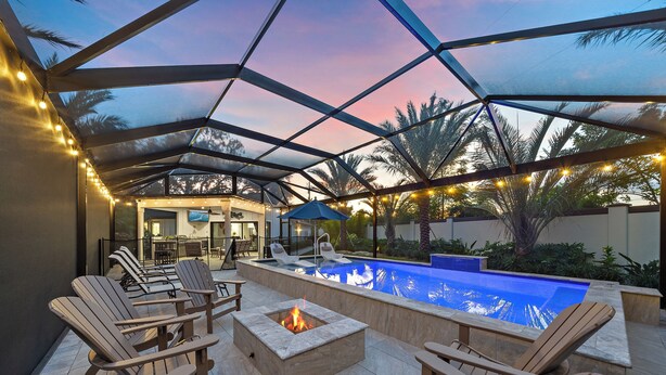 Zona de piscina exterior con sillas de descanso, un hogar para fogata y un patio cubierto con guirnaldas de luces.