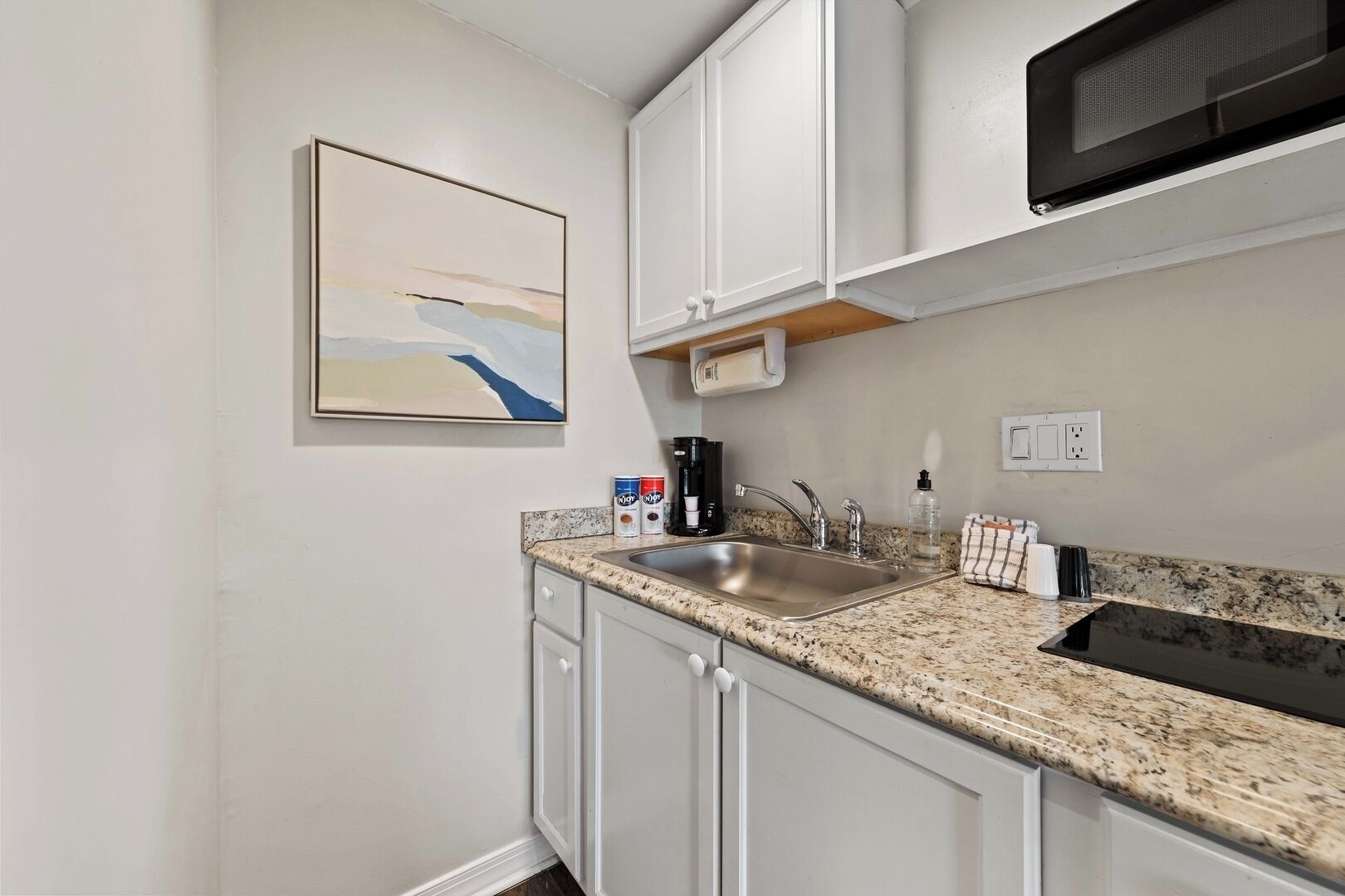 A kitchen with white cabinets, a granite countertop, a stainless steel sink, a microwave, and a coffee maker.