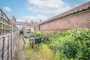 A brick house with a tiled roof, a wooden fence, and a small patio with a table and chairs.