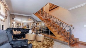 Ein großzügiger Wohnbereich mit einem schwarzen Samtsofa, zwei cremefarbenen Hocker und einem flauschigen Teppich. Eine Holztreppe mit aufwendiger Metallgeländer führt zu einer oberen Ebene.