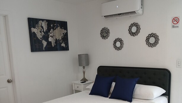 A neatly made bed with a white bedspread and two pillows, a world map artwork on the wall, a wall-mounted air conditioner, and a "no smoking" sign.