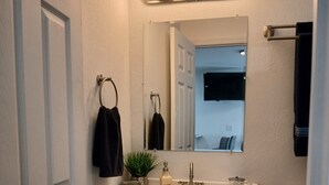 A bathroom with a white vanity, a mirror, a sink with soap and a towel, a toilet, and a trash can.