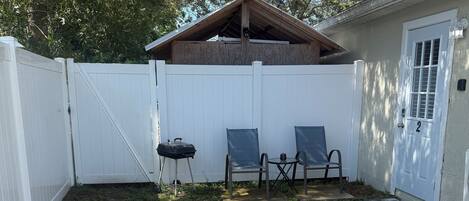 A backyard with a white fence, a covered patio area, and a trash can.