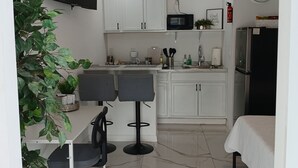 A compact kitchen with white cabinets, a microwave, a clock, and a refrigerator. There are two bar stools and a small table with chairs.