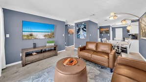 A modern living room with a large flat-screen TV, a brown leather sofa, a wooden coffee table, and a dining area with a white table and chairs. - Poolside Villas 104 near beach sleeps 5 (Destin)