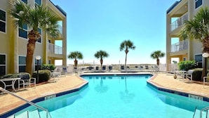 A swimming pool with lounge chairs and palm trees, surrounded by a multi-story building with balconies. - Poolside Villas 104 near beach sleeps 5 (Destin)