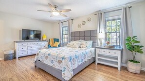 Un dormitorio con cama, cómoda, televisor, ventilador de techo y ventana con cortinas.