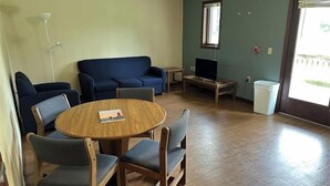 A living room with a round wooden table, four chairs, a blue sofa, a television on a wooden stand, and a window with blinds. - Apartment Centrally Located (Plymouth)