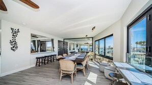 A spacious dining area with a long wooden table and wicker chairs, large windows offering a view of the city, and a modern kitchen visible through an open doorway.