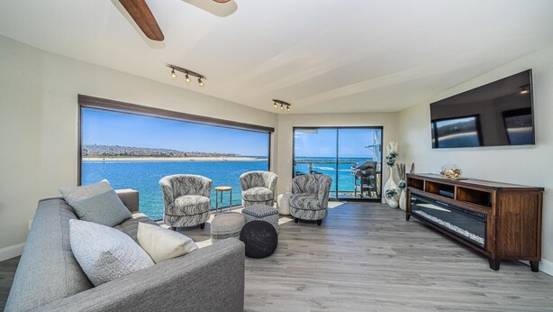 A spacious living room with a large window overlooking a beach, a grey sofa, patterned armchairs, and a wooden entertainment unit.