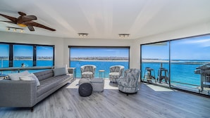 A modern living room with a gray sofa, patterned armchairs, and a ceiling fan. Large windows offer a view of the sea and a distant shoreline.