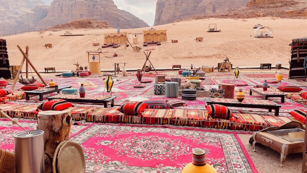 A desert resort with outdoor seating areas covered with colorful rugs and cushions, wooden furniture, and a view of the surrounding rocky landscape. - Luxury Wadi Rum Camp (Wadi Rum)