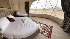 A bedroom with a large bed, a smaller bed, and a view of a desert landscape through a geometric window. - Luxury Wadi Rum Camp (Wadi Rum)