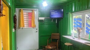 Ein kleiner, abgeschlossener Raum mit grüner Einrichtung, einer weißen Tür, einem an der Wand befestigten Fernseher und einem Fenster mit Blick auf einen Spielplatz.