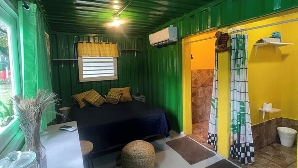 Ein kleines Zimmer mit einem Bett, einem Nachttisch und einem kleinen runden Hocker. Es gibt ein Fenster mit Vorhängen, eine Klimaanlage und ein Badezimmer mit Duschvorhang.