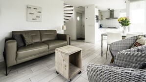 Ein modernes Wohnzimmer mit einer Couch, einem kleinen Holztisch und einem Korbstuhl. Im Hintergrund ist eine Küchenzeile mit einem Esstisch und Stühlen zu sehen.