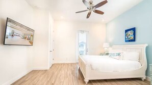 A bedroom with a white bed, a ceiling fan, a framed picture on the wall, and a view of the outside.