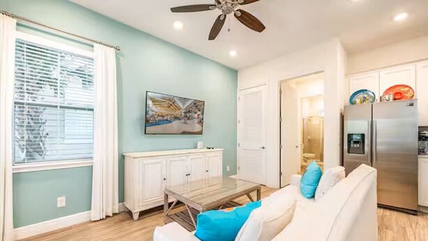 A living room with a white sofa, a coffee table, and a ceiling fan. There is a kitchen area with a refrigerator and cabinets.