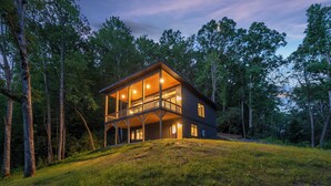 Une cabane en bois à deux étages, dotée de grandes fenêtres et d’un balcon, entourée d’arbres.