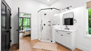 Une salle de bain moderne avec une douche à l’italienne en verre, un lavabo blanc avec un miroir et une vue sur la verdure à l’extérieur.
