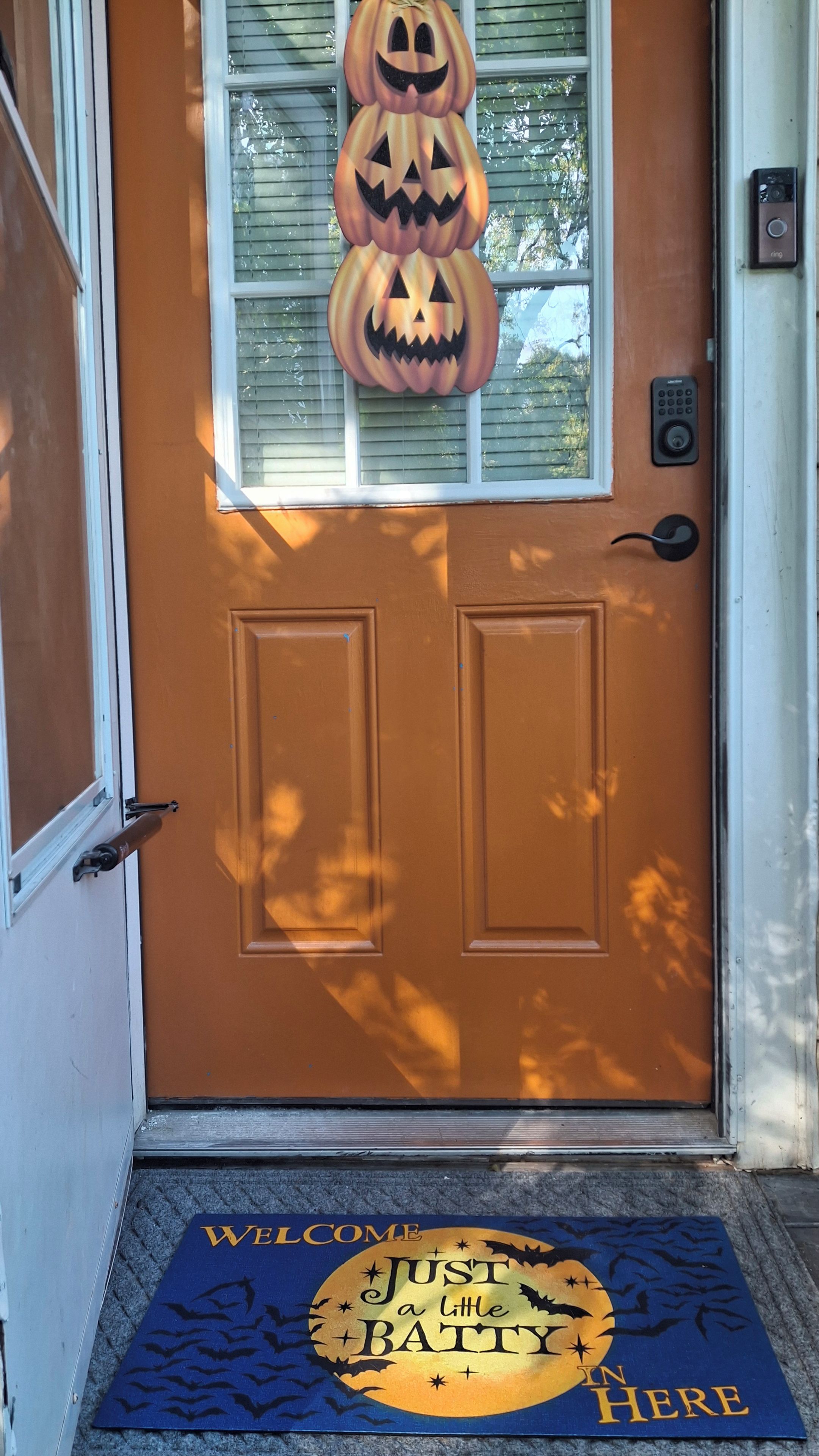 Une porte avec un paillasson sur le thème d’Halloween, une fenêtre décorée d’une citrouille et un carillon.