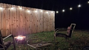 Cerca de madeira com luzes de cordão, uma cadeira de metal, uma mesa pequena e um braseiro.