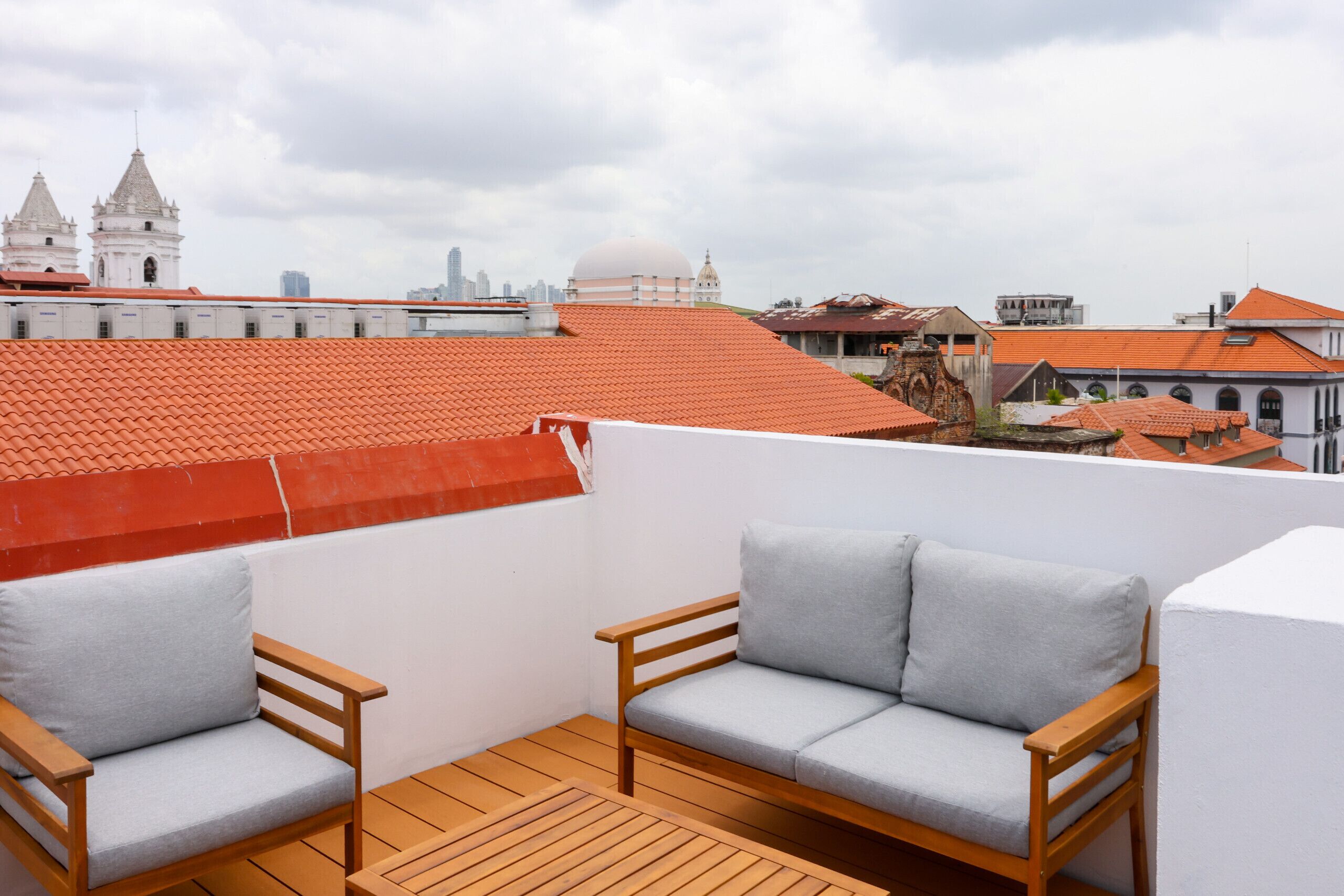 A rooftop terrace with two wooden chairs, a wooden table, and a view of city buildings and a dome structure.