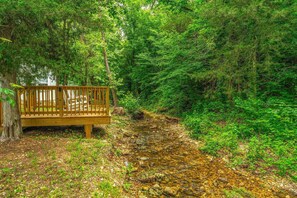 A wooden bench by a forested area with a stream. - Relaxing Wooded Retreat | Pool Access, Near Dogwood Canyon and Thunder Ridge (Blue Eye)
