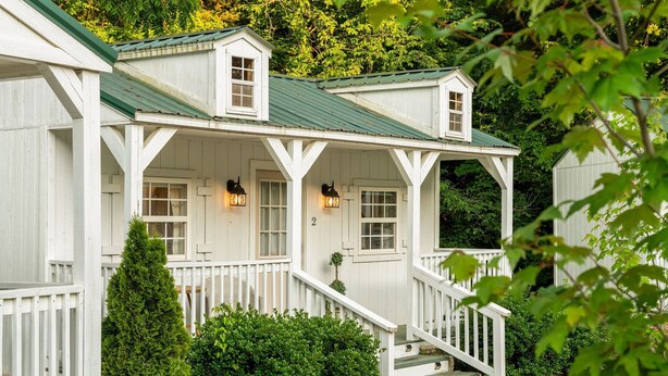 Une maison en bois blanche, avec un toit vert, une véranda dotée de balustrades blanches et un escalier menant à l’entrée.