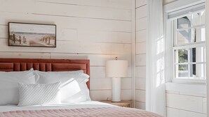 Une chambre à coucher avec une tête de lit en bois, un lit recouvert d’une literie blanche et rose, une lampe de chevet et une fenêtre avec des volets blancs.