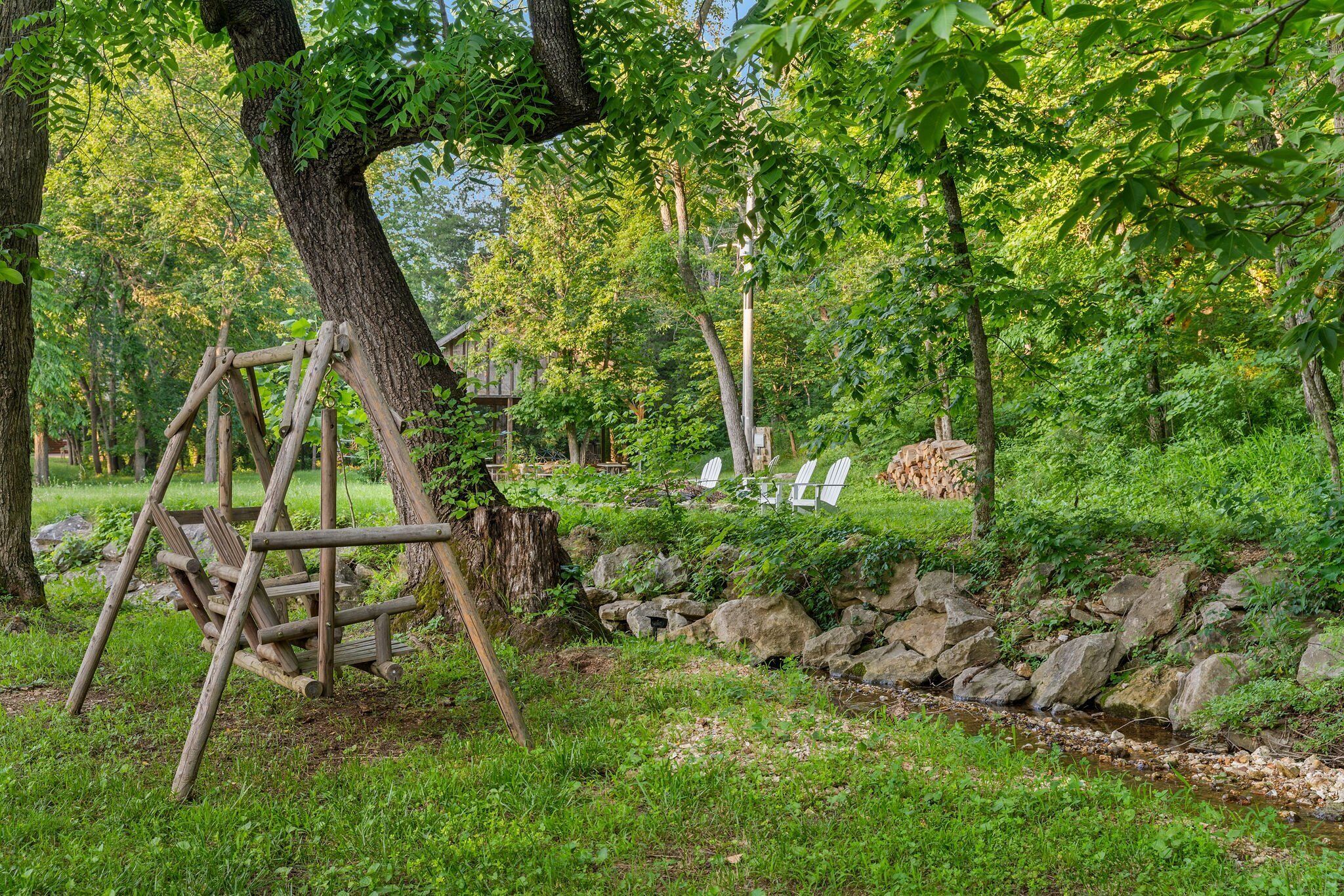 A wooden swing set, a small stream, and a pile of firewood in a lush green park.
