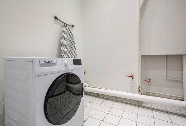 A compact, modern laundry room with a front-loading washing machine, a drying rack, and a white tiled floor.