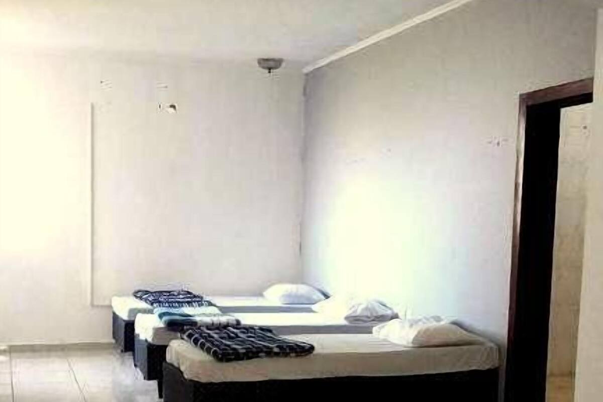 Habitación de hotel con tres camas dispuestas en fila, piso de baldosas y pared blanca lisa.
