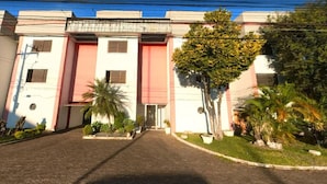 Un edificio con una entrada central, flanqueado por dos alas. Palmeras y arbustos en primer plano.