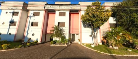 A building with a central entrance, flanked by two wings. There are palm trees and shrubs in the foreground.