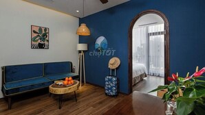 A room with a blue wall, a wooden ceiling fan, a blue sofa, a round table with oranges, a lamp, a suitcase, and a bed with a window.