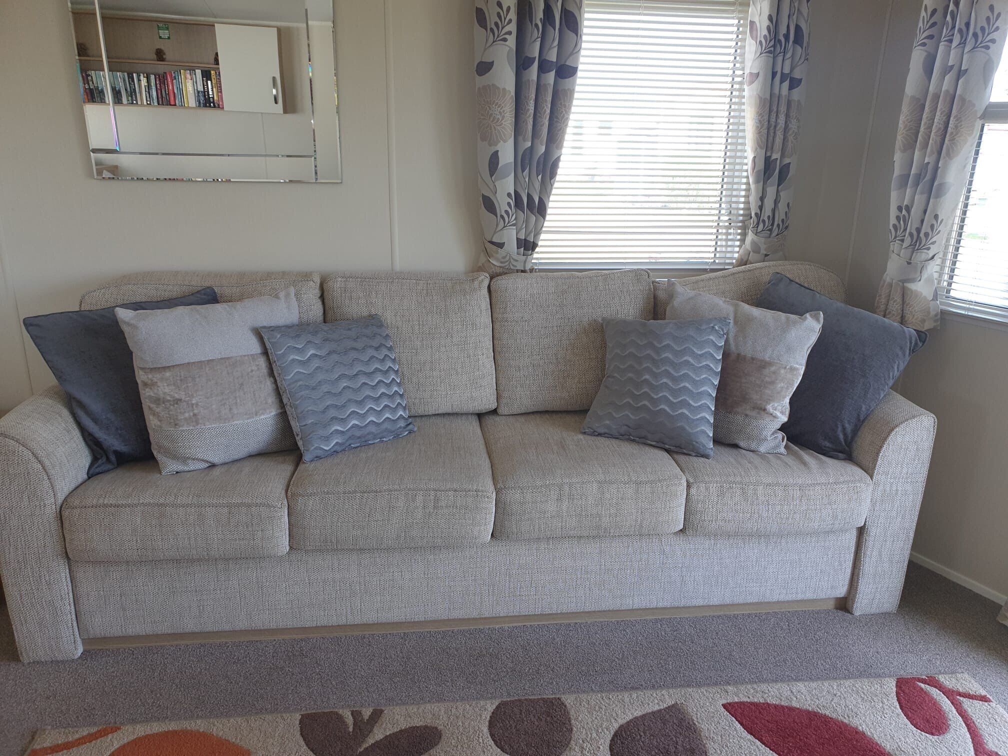 A living room with a grey sofa, patterned cushions, a bookshelf, and a window with curtains.