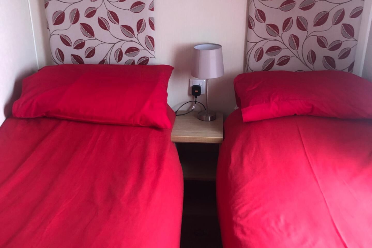 Twee eenpersoonsbedden met rode beddengoed, een bedlampje en een stopcontact op een nachtkastje van hout.