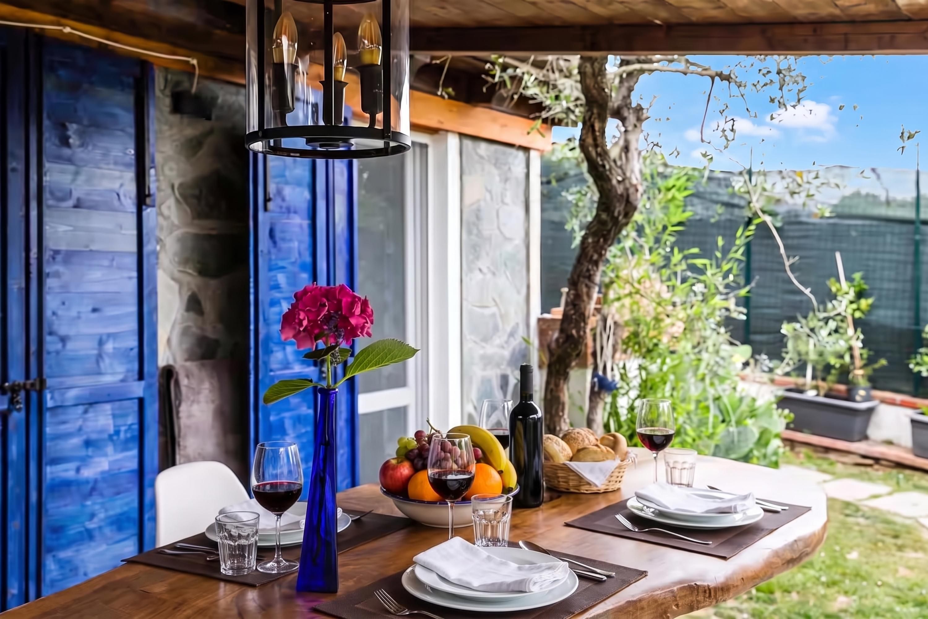 An outdoor dining area with a table set for two, featuring a vase with flowers, a bowl of fruit, wine glasses, and a bottle of wine.