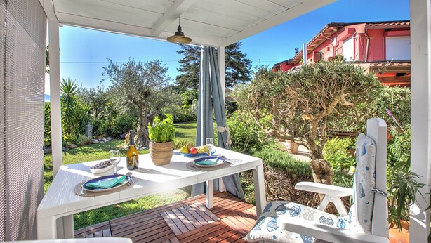 An outdoor patio with a table set for dining, surrounded by greenery and a view of a red building.