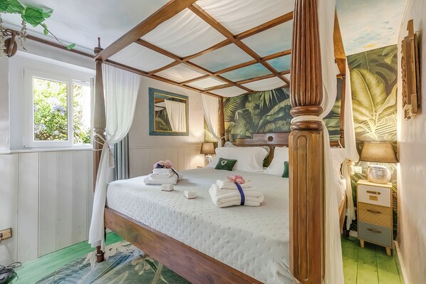 Una cama con dosel, una mesita de noche con lámpara y un cuadro con follaje tropical en la pared.
