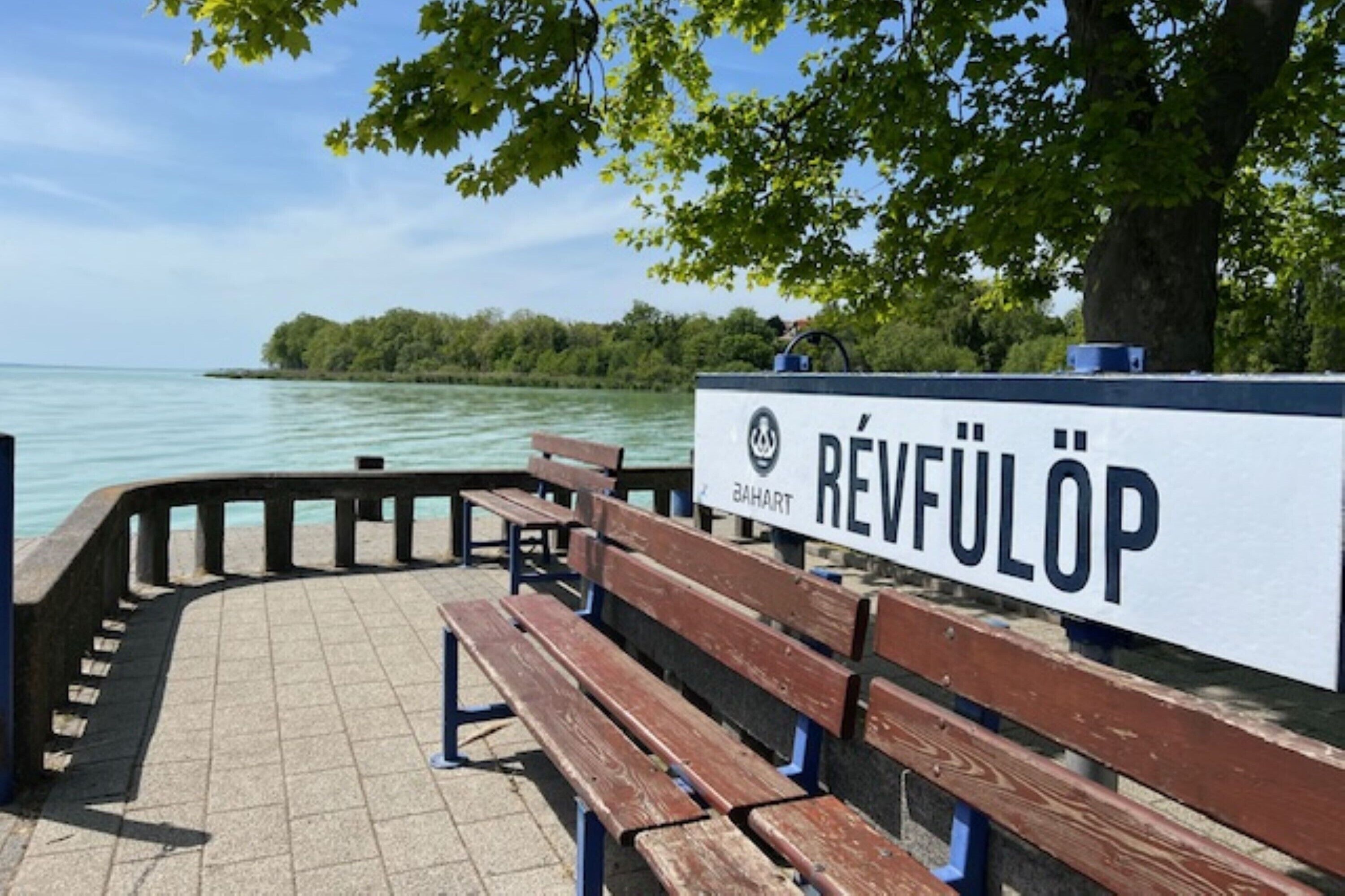 Un banc et une plaque portant le nom « Révfülöp », situés dans un parc, près d’une étendue d’eau.