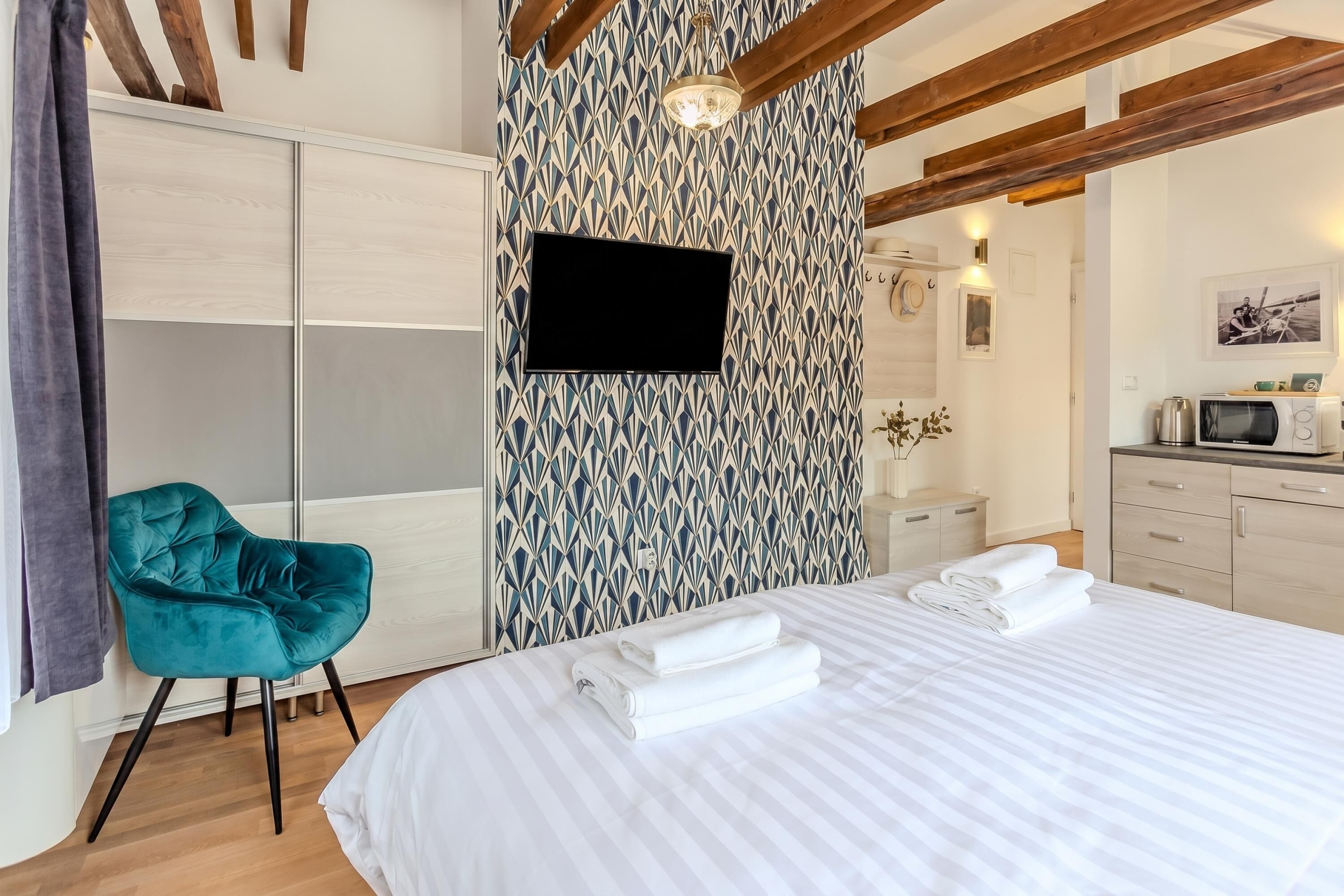 Een hotelkamer met een bed, een blauwgroene stoel, een televisie, een magnetron en een kamer met een patroon op de muur.