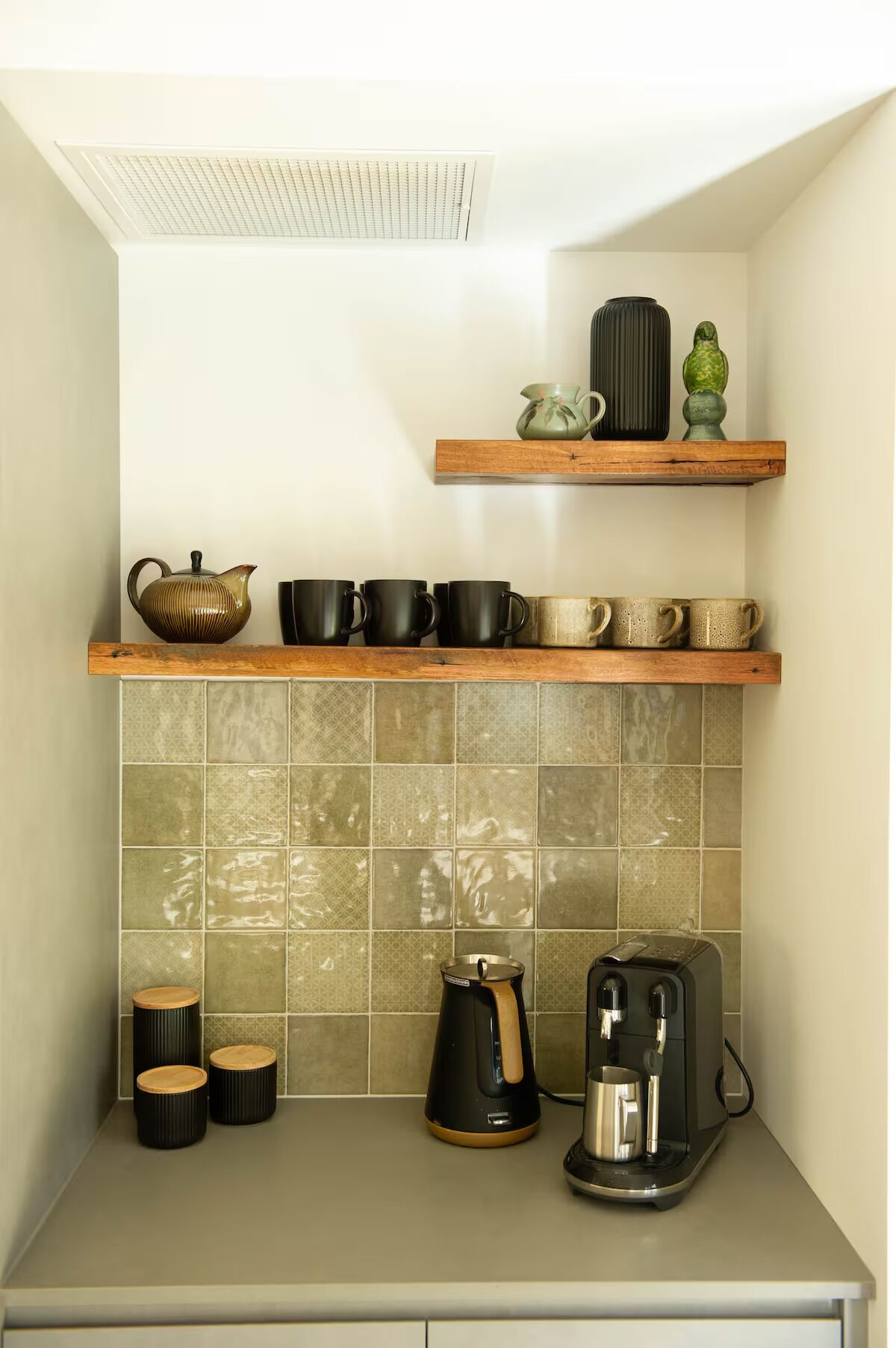 Eine kompakte Küche mit einer Kaffeemaschine, einem Wasserkocher und einem Toaster auf der Arbeitsfläche. Darüber befinden sich zwei Holzböden mit verschiedenen Küchenutensilien.